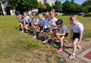 10 dziewczynek stoi na starcie biegu na trawie obok boiska
