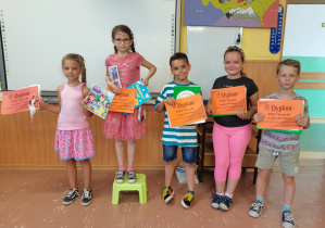 5 stojących przy tablicy dzieci trzyma dyplomy i nagrody za klasowy Konkurs Układania Wyrazów