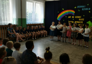 Siedmioro uczniów klasy I czyta wiersze i życzenia stojąc na środku sceny. W tle - zaproszony gość oraz uczniowie Bajkolandii