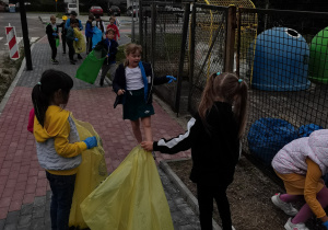Kilkoro dzieci trzyma worki i zbiera śmieci na chodniku przy szkole.