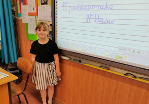 Dziewczynka prowadząca zajęcia stoi przed tablicą przy biurku.