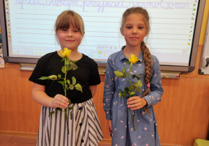Dwie dziewczynki stoją przed tablicą, w rękach trzymają kwiatki.