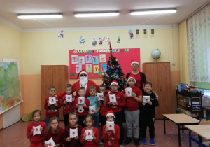Uczniowie z klasy I a w strojach mikołajkowych wraz z wychowawczynią pozują do zdjęcia z Mikołajem.