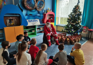 Mikołaj siedzi w otoczeniu dzieci. W tle choinka.