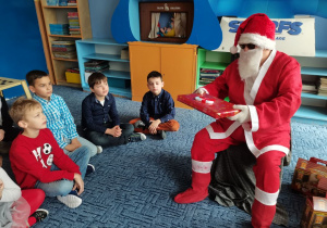 Mikołaj trzyma w ręku prezent. Dzieci obserwują gościa.