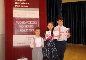 Troje dzieci stoi przy banerze konkursowym i prezentuje dyplomy.