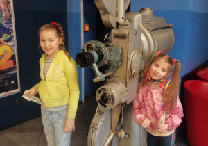 2 dziewczynki pozują do zdjęcia w holu kina przy dużym projektorze.