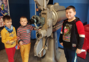 3 chłopców pozuje do zdjęcia w holu kina przy dużym projektorze.