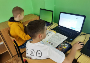 2 chłopców siedzi przy laptopach w pracowni komputerowej.
