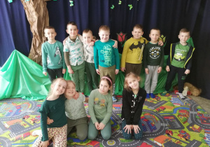 Dzieci w zielonych strojach stoją na tle wiosennej dekoracji. 7 chłopców stoi, a 4 dziewczynki klęczą przed nimi.