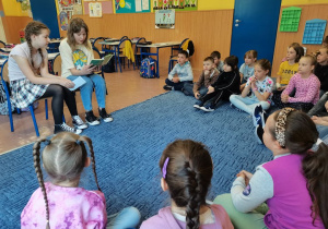 Dzieci siedzą na dywanie. 2 uczennice z klasy V czytają im książkę.