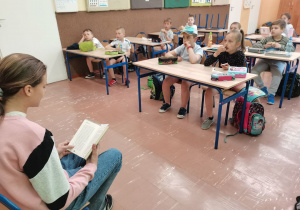 Dzieci siedzą w ławkach. Uczennica z klasy V czyta im książkę.