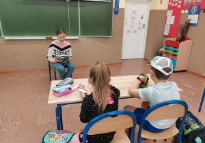 Dzieci siedzą w ławkach. Uczennica z klasy V czyta im książkę.