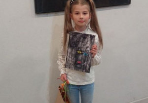 Dziewczynka stoi przed wystawą prac, trzyma dyplom i nagrodę.