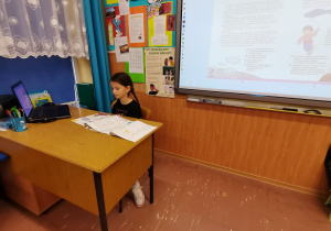 Uczennica siedzi za biurkiem nauczyciela.
