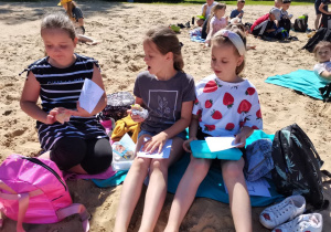 3 dziewczynki siedzą na piasku i jedzą kanapki.