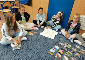 Uczniowie siedzą na dywanie, opowiadają o wakacjach i prezentują pamiątki.