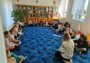 Dzieci wraz z panią bibliotekarką siedzą w kręgu na dywanie w czytelni.