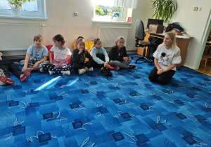Dzieci wraz z panią bibliotekarką siedzą w kręgu na dywanie w czytelni.