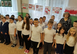 Uczniowie w strojach galowych biorą udział w akcji śpiewania hymnu państwowego.