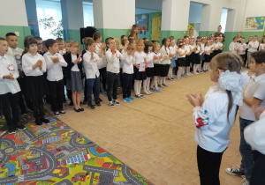 Uczniowie w strojach galowych biorą udział w akcji śpiewania hymnu państwowego.