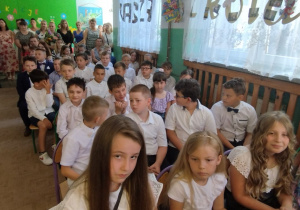 Uczniowie słuchają siedząc w holu szkoły